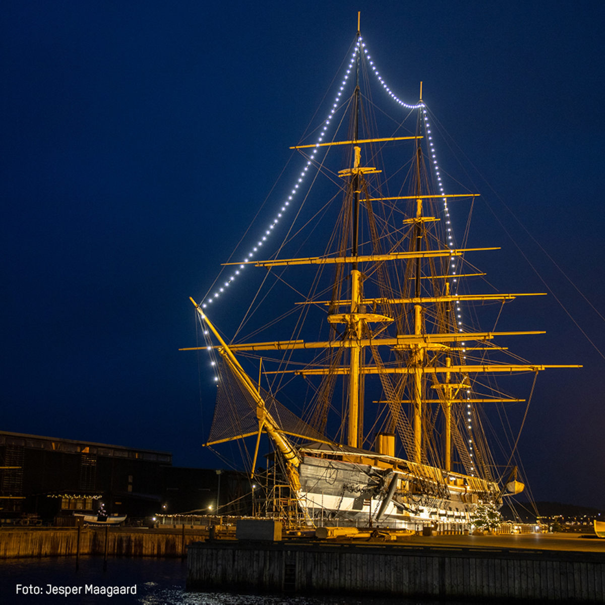 Historisk sejlskib belyst om natten i havn med mastetråd oplyst mod den mørke himmel.
