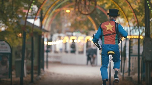 Cirkusartist på ethjulet cykel iført farverigt kostume med stjerne på ryggen cykler gennem parkens lyshimmel.