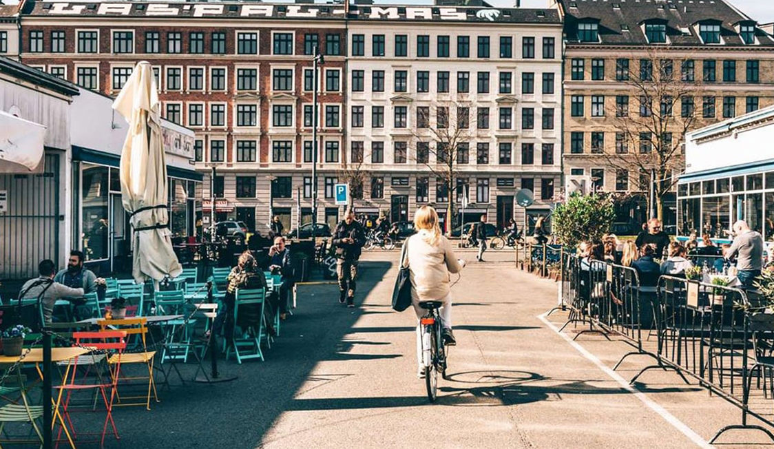 Cyklist på huvudgatan i Kodbyen, omgiven av uteserveringar med människor som njuter av soligt väder.