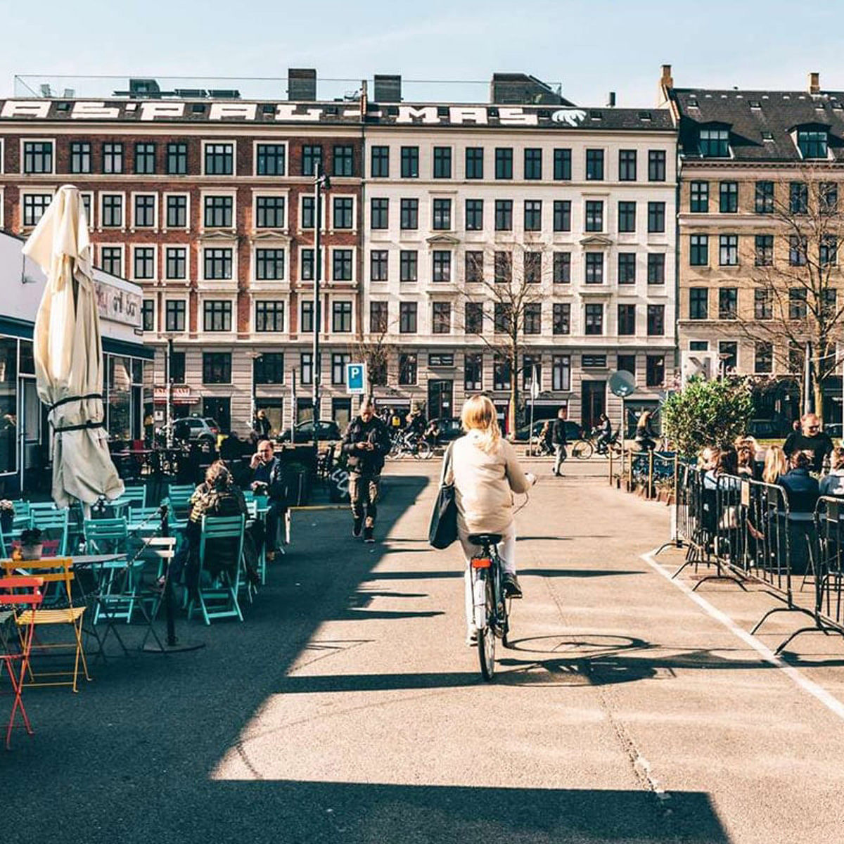 Cyklist på huvudgatan i Kodbyen, omgiven av uteserveringar med människor som njuter av soligt väder.
