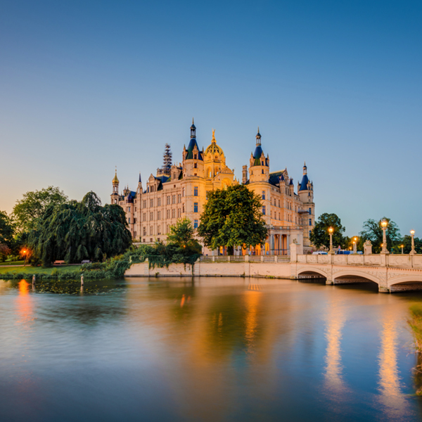 Slottet Schwerin i kvällsljus, omgiven av vatten och grönska, med bro i förgrunden.