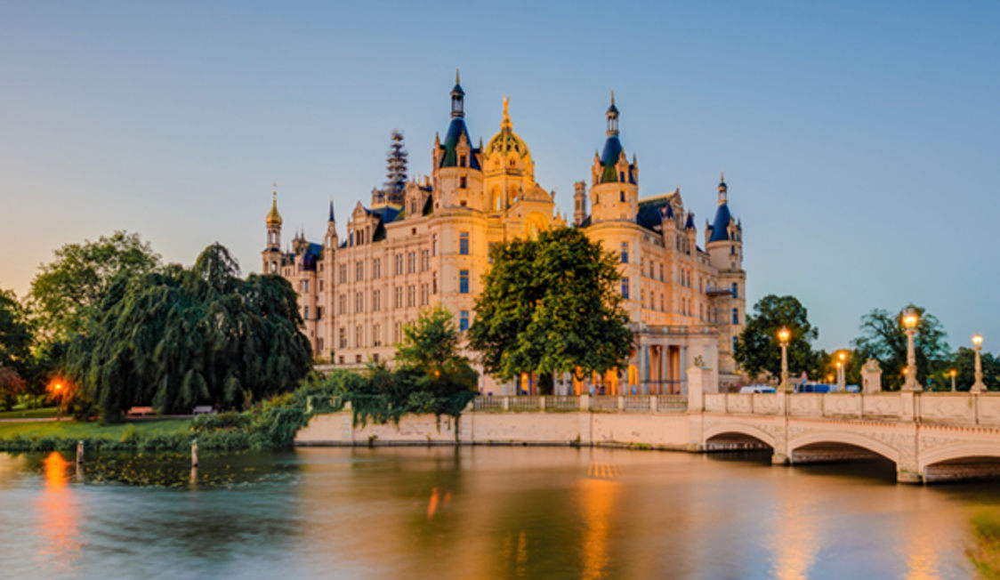 Slottet Schwerin i kvällsljus, omgiven av vatten och grönska, med bro i förgrunden.