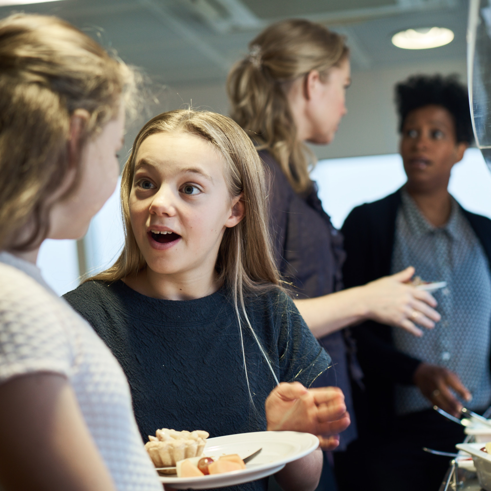 Ung pige i grå bluse udtrykker overraskelse, mens hun taler med en anden person indendørs.