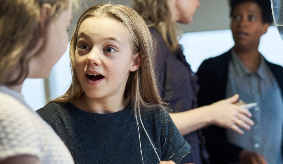 Ung flicka i grå tröja uttrycker förvåning medan hon pratar med en annan person inomhus.