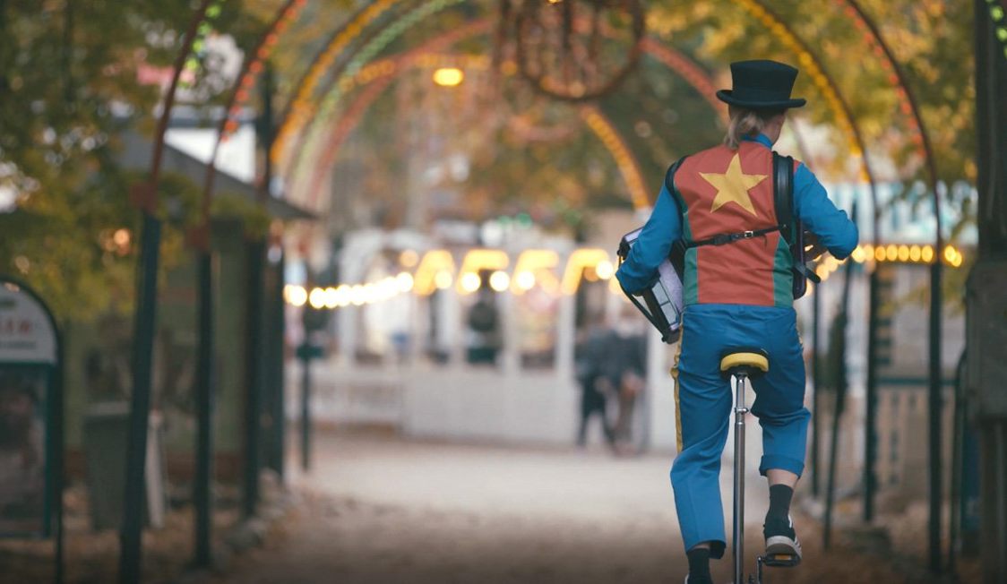 Cirkusartist på enhjulet cykel med farverig kostume og trommer, under lysbue i en forlystelsespark.