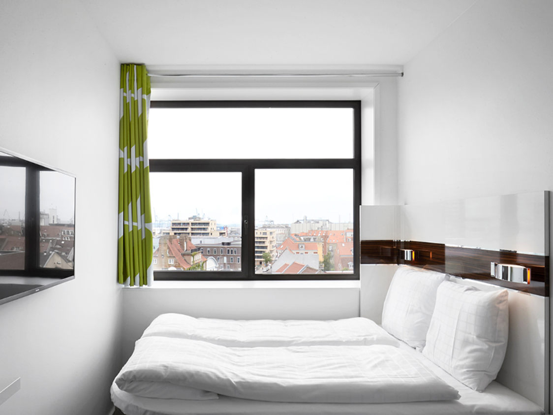 Standard hotelværelse med hvid seng, grønne gardiner og udsigt over byens tage fra stort vindue.