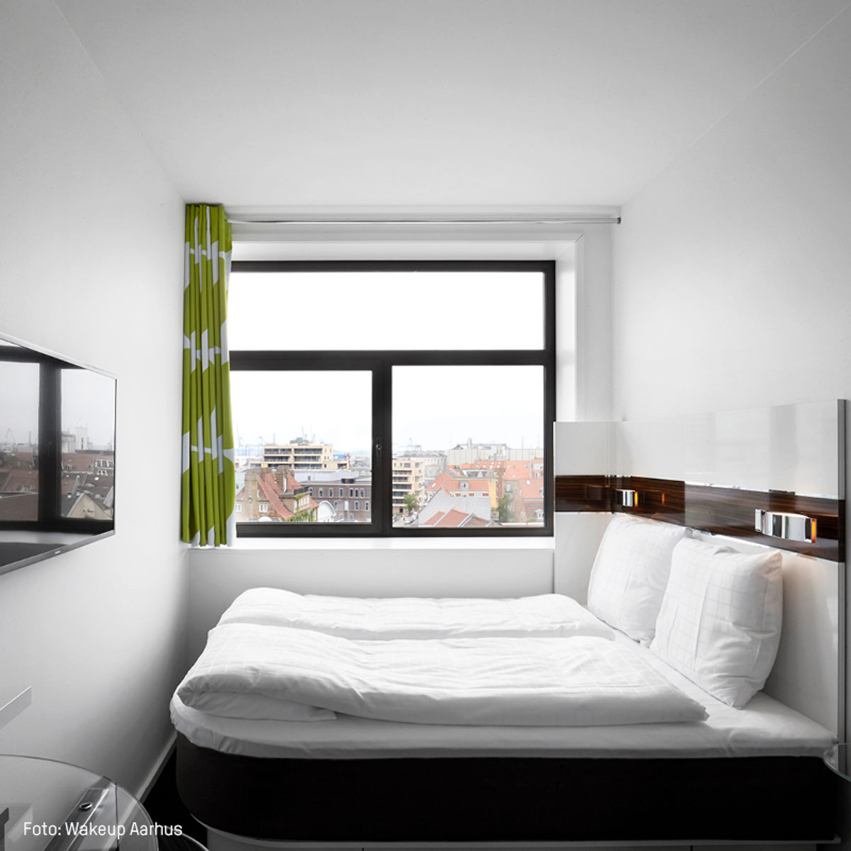 Standard hotelværelse med hvid seng, grønne gardiner og udsigt over byens tage fra stort vindue.