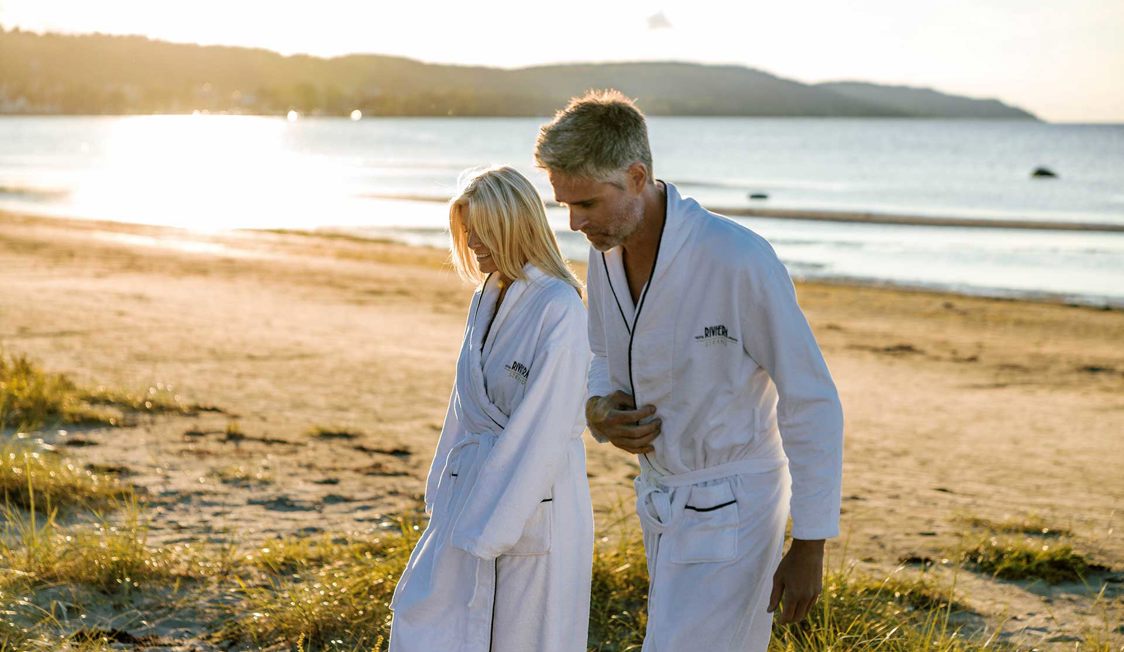 To personer i hvide badekåber går på en solrig strand ved vandet.
