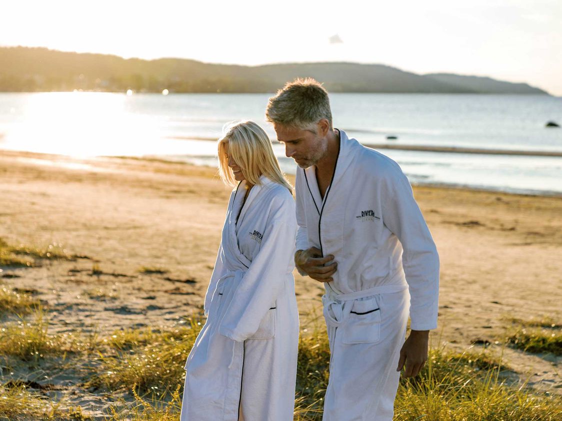 To personer i hvide badekåber går på en solrig strand ved vandet.