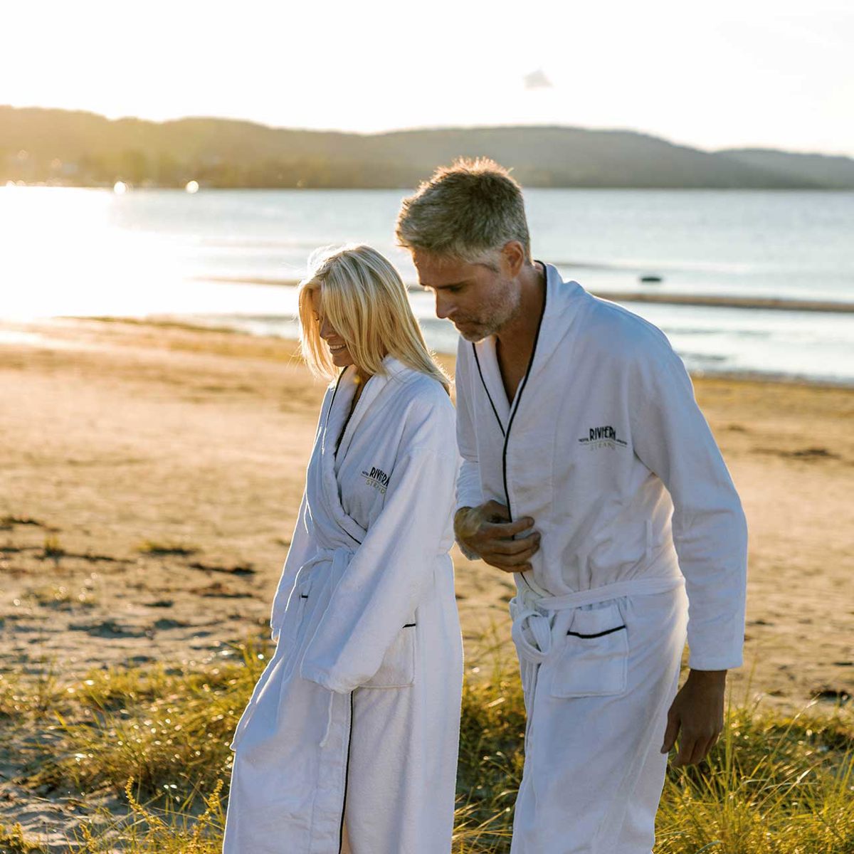 To personer i hvide badekåber går på en solrig strand ved vandet.